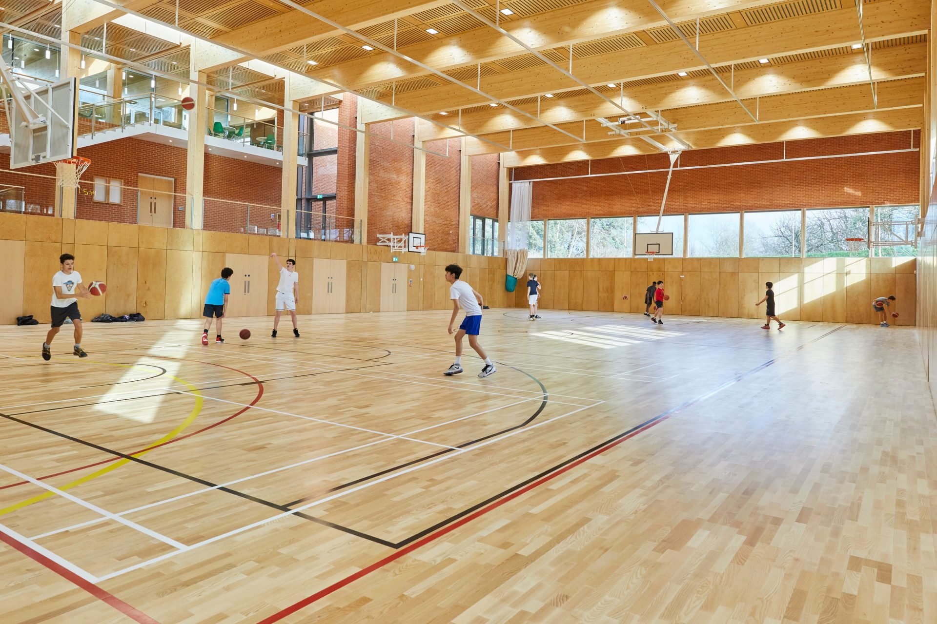 The mass-timber interior of the sports hall at Eton College Sports & Aquatic Centre, with boys playing basketball.